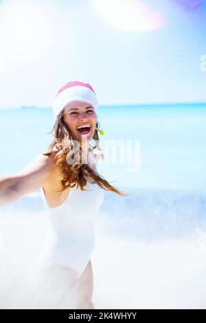 bonne femme moderne d'âge moyen en vêtements de plage blancs avec chapeau de noël rayé ayant du plaisir à la plage. Banque D'Images