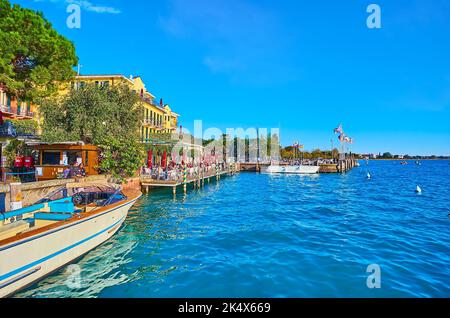 La surface bleu vif ondulée du lac de Garde à partir de l'embarcadère de ferry de Sirmione avec une vue sur le navire de départ et la ligne de drapeaux ondulés de différents Banque D'Images