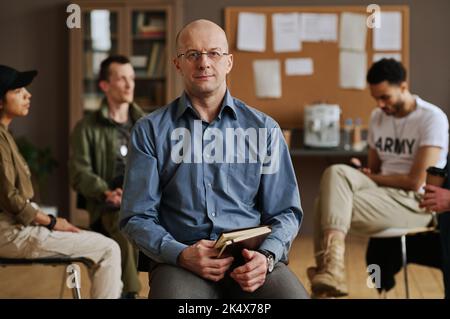 Conseiller ou psychologue homme mature et confiant avec ordinateur portable regardant la caméra tout en étant assis contre un groupe de jeunes patients interculturels Banque D'Images