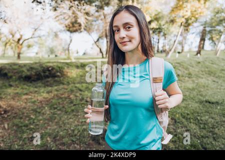 Adolescente portant une chemise turquoise et un sac à dos rose, tenant une bouteille de verre avec de l'eau dans la nature Banque D'Images