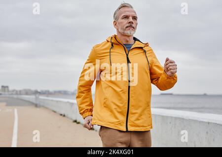 Taille vers le haut portrait d'un beau homme mature qui court par rivière contre le ciel gris et porte une rainjaquette jaune, espace de copie Banque D'Images