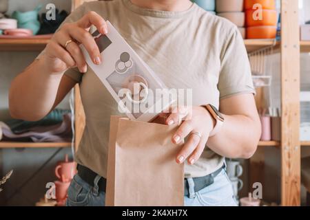 Photo rognée des mains d'une femme d'affaires qui s'emboîte et prépare le colis pour livraison à l'acheteur. Femme met la sucette dans un sac en papier kraft. Petite entreprise Banque D'Images