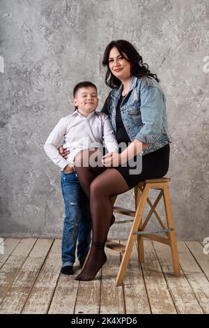 Une mère en robe noire tient son fils dans les bras et s'assoit sur une chaise haute. Une femme et un bébé posent dans un studio isolé sur un fond gris et un Banque D'Images