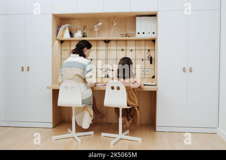 Une jeune mère aide sa petite fille mignonne à faire ses devoirs. Assis au bureau. Vue de derrière. Banque D'Images