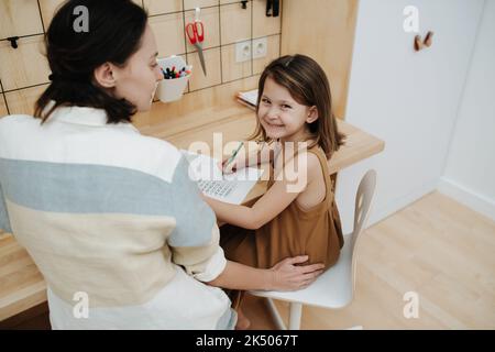 Une fille gcheeky regardant l'appareil photo et sa mère faisant ses devoirs ensemble. Assis au bureau. Vue de derrière. Banque D'Images