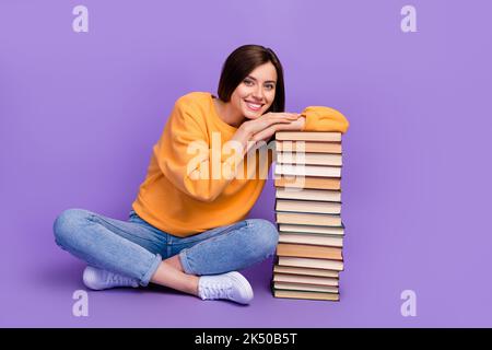 Pleine longueur de corps photo de jeune fille positive rêveuse assis jambes croisées adorable toucher sa maison bibliothèque pile livres nerd isolé sur la couleur pourpre Banque D'Images