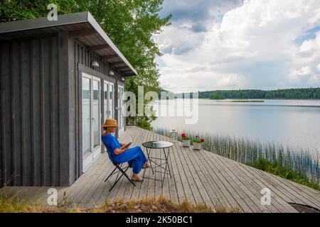 Une femme âgée assise lisant sur une terrasse en bois à l'extérieur d'une maison de plage à côté d'un lac Banque D'Images