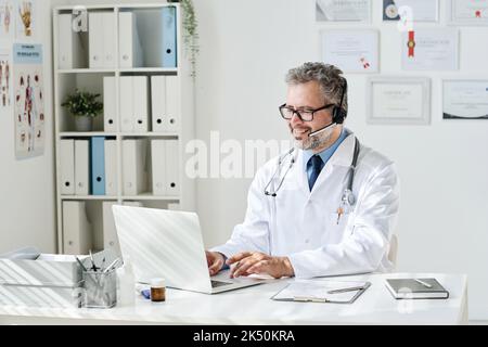 Médecin mature dans les écouteurs assis à la table devant l'ordinateur et ayant la consultation en ligne Banque D'Images