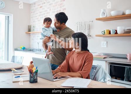 Famille, bébé et le syndrome de Down alors que la mère travaille en ligne avec un ordinateur portable dans la cuisine. Maman, père et enfant jouent tandis que la mère utilise l'ordinateur pour l'apprentissage Banque D'Images