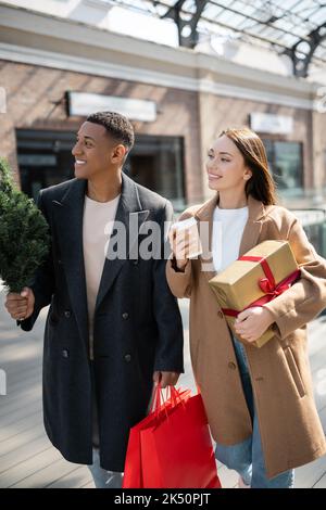 couple interracial en manteaux tenant des cadeaux du nouvel an et petit arbre de noël tout en regardant loin sur la rue de la ville, image de stock Banque D'Images