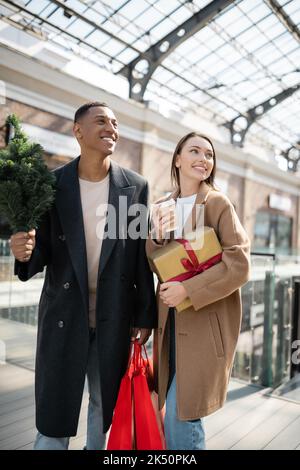 couple interracial élégant avec des cadeaux du nouvel an et petit arbre de noël souriant près du centre commercial flou, image de stock Banque D'Images