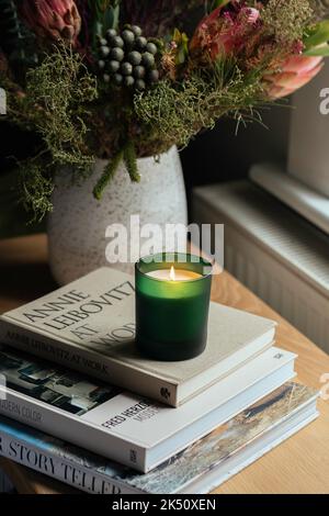 Bougie de soja dans un pot en verre vert assis sur une pile de livres dans la chambre parmi les fleurs automnales Banque D'Images