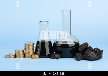 Pétrole brut dans des béchers en verre et des morceaux de charbon dur noir avec des piles de pièces sur fond bleu, concept de prix des combustibles fossiles Banque D'Images