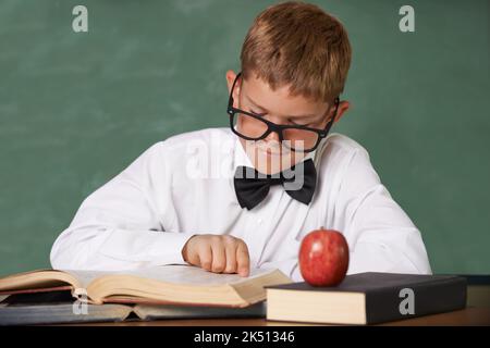 Il aime lire. Un jeune garçon portant des lunettes et un noeud papillon se concentrant sur sa lecture avec une pomme rouge sur la table en face de lui. Banque D'Images