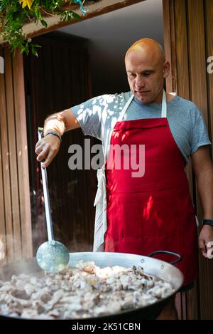 Chef masculin adulte concentré en tablier remuant les ingrédients dans une grande casserole en métal tout en préparant la paella traditionnelle dans le jardin le jour ensoleillé Banque D'Images