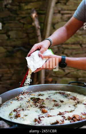 Cultivez le chef anonyme en ajoutant du riz dans une grande casserole en métal avec du poulet et des haricots tout en préparant la paella traditionnelle dans la campagne Banque D'Images