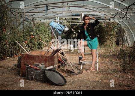 Tout le corps d'une agricultrice assise sur une échelle près de divers instruments de jardinage dans une serre à la campagne et en regardant loin Banque D'Images