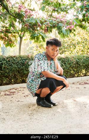 Personne asiatique non binaire à corps entier dans une chemise et une jupe élégantes penchée à la main et regardant la caméra tout en étant assis sur des haunches le jour d'été ensoleillé en pa Banque D'Images