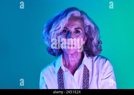 Portrait d'une femme âgée confiante avec des cheveux courts en argent et en chemise blanche regardant l'appareil photo dans les lumières vertes et violettes Banque D'Images