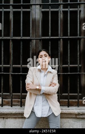 Femme hispanique adulte confiante aux cheveux foncés et aux bras croisés dans une tenue élégante et décontractée, à proximité d'un bâtiment vieillissant avec barres de fenêtre en métal et à l'aspect Banque D'Images