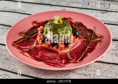 Au-dessus de la salade de tomates avec burrata et des tranches de prosciutto garnies d'olives noires et de sauce pesto et servies sur une assiette sur une table en bois Banque D'Images