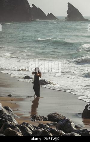 jolie et tatouée femme en robe noire ajustement des cheveux tout en se tenant sur la plage de sable au portugal, image de stock Banque D'Images