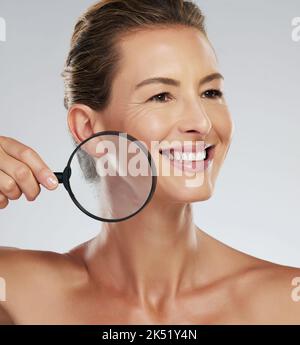 Soin de la peau, beauté et femme mûre avec loupe pour l'inspection du visage sur un fond gris maquette studio. Modèle plus âgé avec sourire pendant Banque D'Images