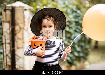 Petite fille mignonne en costume de sorcière tenant le ballon, stand à proximité jack-o-lanterne seau de citrouille avec bonbons et bonbons. Trick d'enfant ou de traitement dans Hallow Banque D'Images