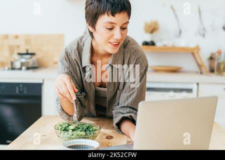 Une jeune femme attirante dans la cuisine mange une salade verte saine et travaille sur un ordinateur portable. Banque D'Images