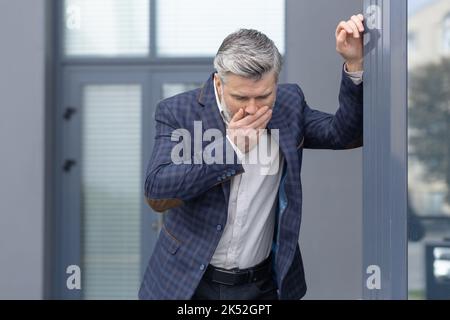Homme d'affaires senior aux cheveux gris malade à l'extérieur du bâtiment moderne de bureau, homme mature dans un costume d'affaires vomissant, patron empoisonné avec des maux d'estomac. Banque D'Images