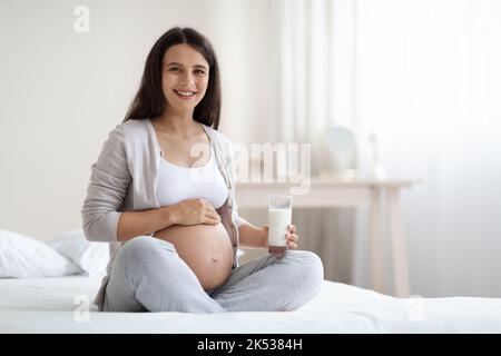 Belle femme enceinte assise sur le lit, tenant un verre de lait Banque D'Images