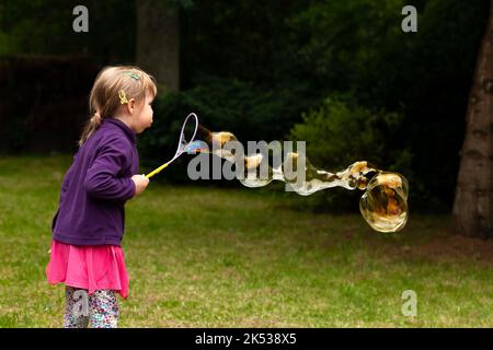 Joyeux enfant d'âge scolaire soufflant d'énormes bulles de savon à l'extérieur, jeune fille caucasienne faisant une grande bulle de savon à l'extérieur dans le parc, seul. Ayant Banque D'Images