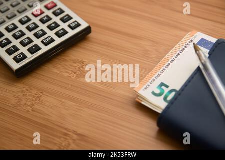 Image d'une calculatrice à côté d'un portefeuille avec des billets en euros sur une table en bois. Image avec espace de copie Banque D'Images