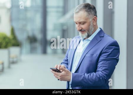 Un beau homme de haut niveau aux cheveux gris en costume se tient dans la rue, tient un téléphone dans sa main, compose un numéro, appelle quelqu'un, sourit. Banque D'Images