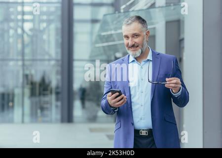 Portrait senior beau gris-cheveux enseignant, professeur, conférencier se tient dans un costume dans un campus moderne, tient un téléphone et des lunettes. Il regarde la caméra, sourit. Banque D'Images