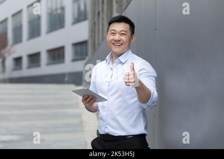 Portrait d'un jeune homme asiatique beau dans une chemise blanche debout dans la rue près d'un bâtiment moderne. Tient une tablette dans sa main, types, fonctionne. Affiche super. Il regarde la caméra, sourit. Banque D'Images