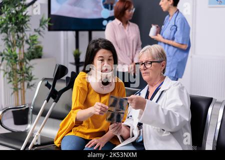 Spécialiste senior consultant le patient avec col cervical regardant l'examen de radiographie médicale pendant la visite de contrôle dans la salle d'attente de l'hôpital. Femme asiatique avec douleur physique et blessure ayant rendez-vous. Banque D'Images