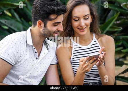 Jeune couple adulte regardant le contenu de réseau social sur l'écran de smartphone - homme et femme utilisant le téléphone assis à l'extérieur et tenant le téléphone portable - les gens an Banque D'Images