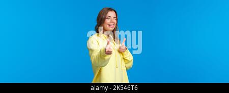 Bonjour à tous. Jolie fille européenne moderne et insouciante dans un sweat à capuche jaune, enfilez et faites des gestes de pistolet à l'appareil photo pour dire salut, sans façons Banque D'Images