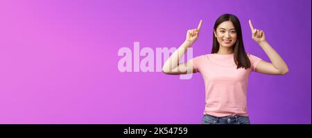 Adorable femme asiatique aux cheveux sombres et affirmée porter un t-shirt rose lever les mains, montrer la promotion, pointer les doigts vers le haut, sourire, inviter utiliser le lien, essayez Banque D'Images