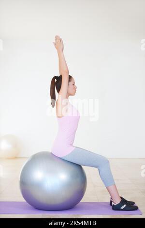 Travailler sur son noyau. Photo de profil d'une jeune femme assise sur une balle d'exercice et étirant ses bras au-dessus de sa tête. Banque D'Images