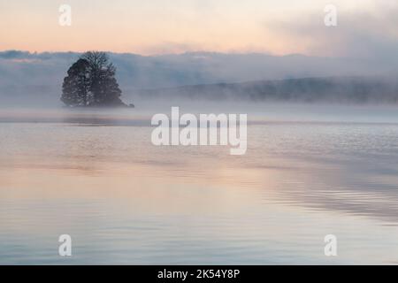Lumière du soleil pâle à l'aube se reflétant sur un lac brumeux avec une île Banque D'Images