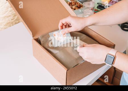 Photo rognée des mains de femme d'affaires emballage, préparation du colis pour livraison à l'acheteur, mise des cartes postales dans la boîte. Propriétaire de petite entreprise Banque D'Images