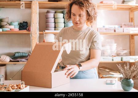 Femme blonde souriante emballage et emballage expédition, plats de bébé en silicone dans une boîte de livraison en carton. Perles de bois brunes dans un récipient à proximité Banque D'Images
