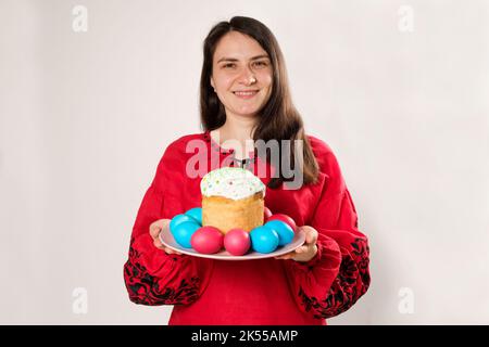 Une femme ukrainienne vêtue de vêtements nationaux rouges brodés tient un gâteau de Pâques et peint des oeufs et des sourires sur un fond blanc Banque D'Images