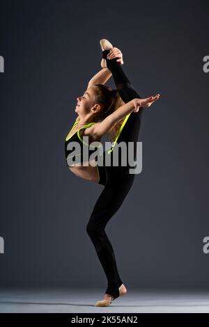 Jeune gymnaste. Une adolescente dans un survêtement est engagée dans la gymnastique, faisant des exercices d'étirement sur un fond gris. Photo de haute qualité Banque D'Images