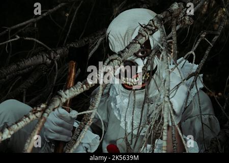 gros plan d'un clown maléfique créepy vu à travers les branches d'un arbre dans les bois la nuit, portant un costume gris avec une robe blanche, avec une hache dans son Banque D'Images