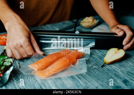 gros plan d'un jeune homme qui empoche sous vide deux morceaux de saumon dans un sac sous vide, sur une table grise rustique Banque D'Images