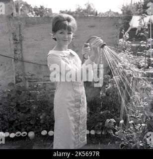 1964, historique, l'été et à l'extérieur dans un jardin, une jeune femme dans une robe arroser ses plantes, en utilisant une pipe à hope avec un spray, Angleterre, Royaume-Uni. Banque D'Images