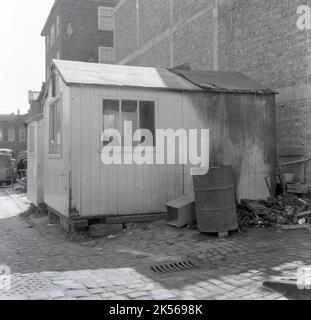 1960s, vue latérale historique d'une cabane en bois d'ouvrier, avec fenêtres, empilées sur des briques sur une zone pavée, Angleterre, Royaume-Uni. À l'extérieur d'un canon métallique utilisé pour brûler des montagnes russes, illustré par l'arrière de la hutte ayant été singué ou sorché. Ces huttes ont été utilisées pour faire une tasse de thé, prendre un repos, changer de vêtements, tenir des outils et une fois un site commun sur les chantiers de construction, les chemins de fer et les centres-villes. Banque D'Images